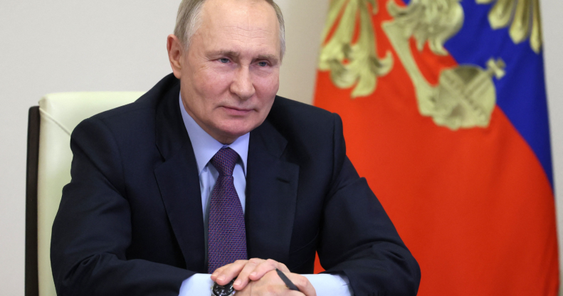 Russian president Vladimir Putin sits at a desk next to a flag with a pen in his hand.