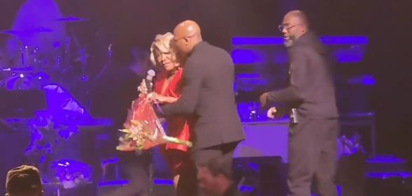 Patti LaBelle, in a red dress, is escorted off stage by two men in black suits, as the rest of the band is escorted off in the background.