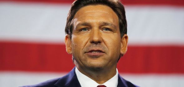 Florida governor Ron DeSantis, wearing a suit and tie, stands in front of the red and white stripes of the US flag