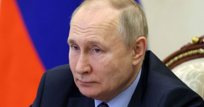 Vladimir Putin stares at someone off camera while sitting in front of a Russian flag after signing an extended LGBTQ+ 'propaganda' bill into law