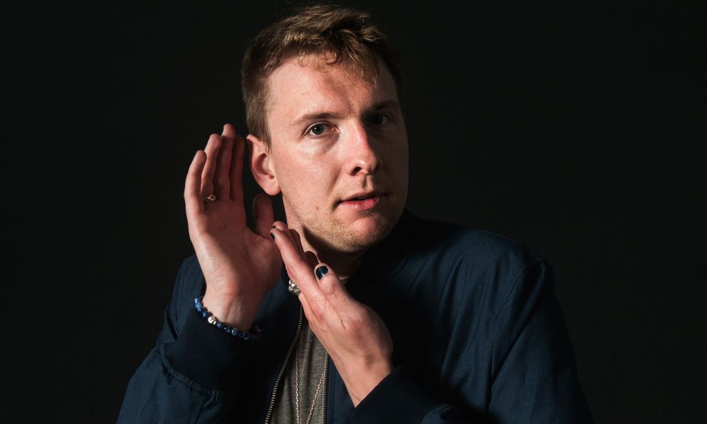 Joe Lycett poses with his hands near his face as he stares towards the camera
