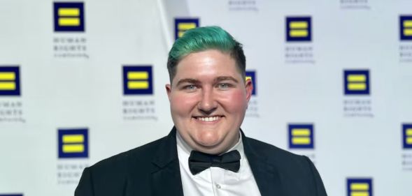 Henry Berg-Brousseau, a trans rights advocate, smiles at the camera while posing in front of logos for the Human Rights Campaign