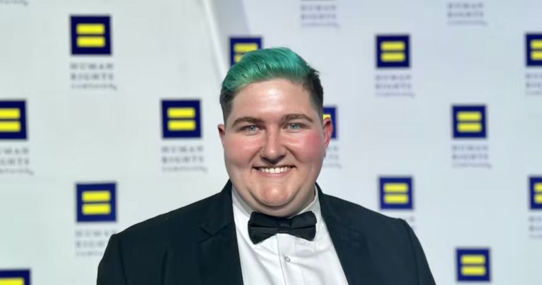 Henry Berg-Brousseau, a trans rights advocate, smiles at the camera while posing in front of logos for the Human Rights Campaign