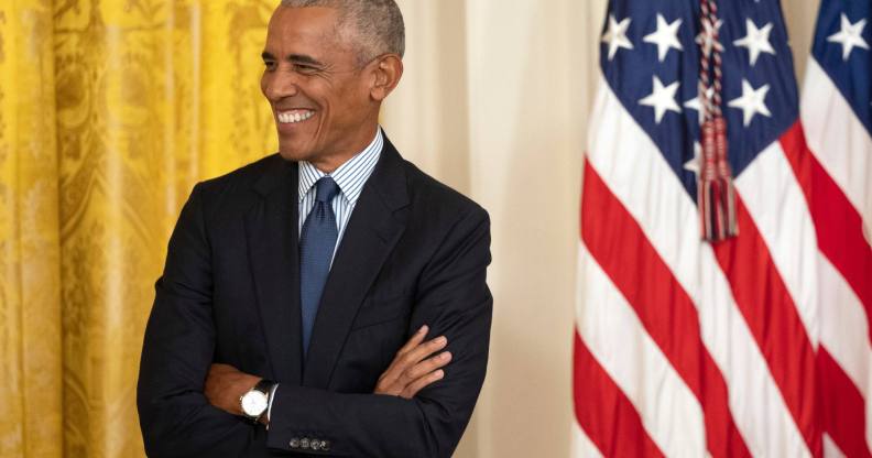 Former president Barack Obama smiles as he wears a suit with his arms crossed. He is standing in front of a red, white and blue American flag