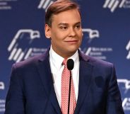 George Santos wears a suit and tie while speaking into a microphone at a conference