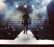 A picture shows a boxer from the back entering a boxing ring - there are crowds of people sitting either side of him