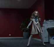 A still from horror film M3GAN showing the robot doll dancing in an office corridor that has red walls and a grey carpet. To the right of the M3GAN robot is a photocopying machine and a small table