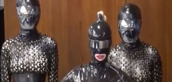A screengrab from Twitter shows three people dressed in black latex, head-to-toe body suits standing in front of a wall
