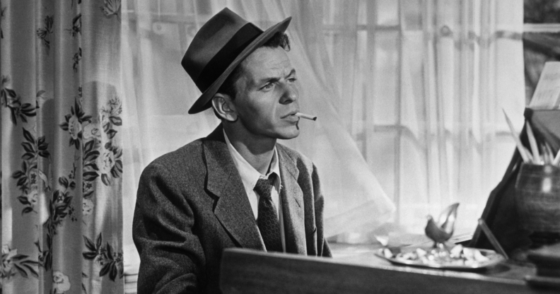 Black and White photo of Frank Sinatra sitting at a piano smoking a cigarette wearing a suit and tie.