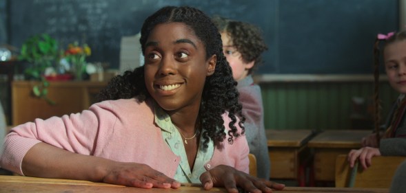 A still from Netflix's Matilda: The Musical showing actor Lashana Lynch as Miss Honey wearing a turquoise shirt and pink cardigan sitting a desk in a classroom