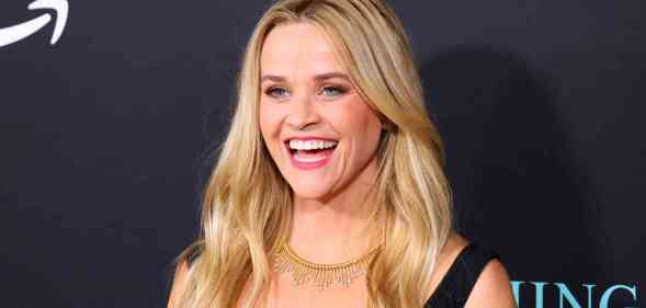 A headshot of actor Reese Witherspoon wearing a black top and smiling as she has her photo taken at a red carpet event