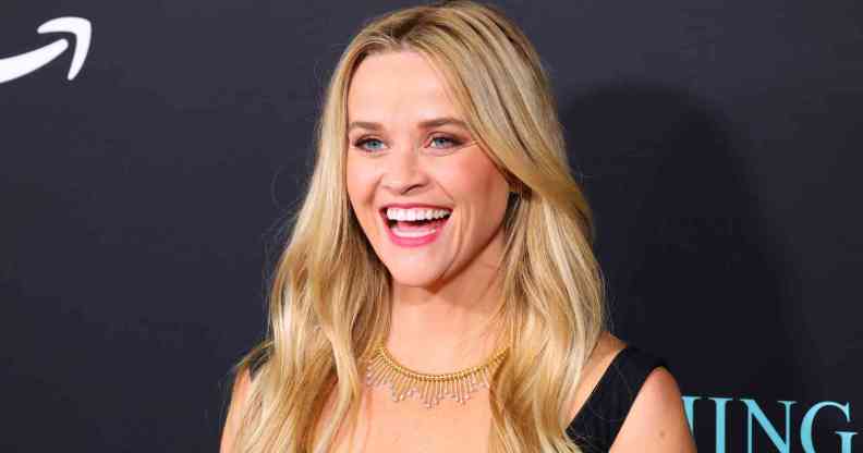 A headshot of actor Reese Witherspoon wearing a black top and smiling as she has her photo taken at a red carpet event