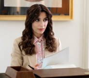 A still from HBO's The White Lotus shows actor Sabrina Impacciatore as Valentina dressed in a cream suit jacket and pink shirt standing behind a reception desk holding some papers.