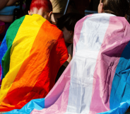 Two people sitting side by side, one in a rainbow flag and one in a trans flag