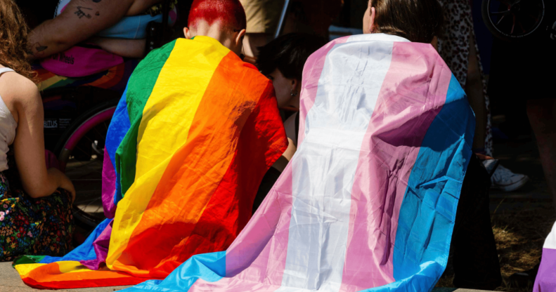 Two people sitting side by side, one in a rainbow flag and one in a trans flag