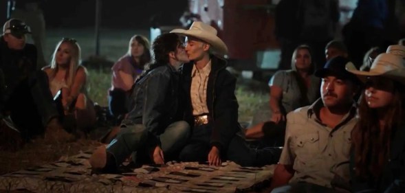 Still from TV series Yellowstone showing actor Lilli Kay as Clara wearing a denim jacket and jeans kissing a woman wearing a black coat, white shirt and white cowboy hat