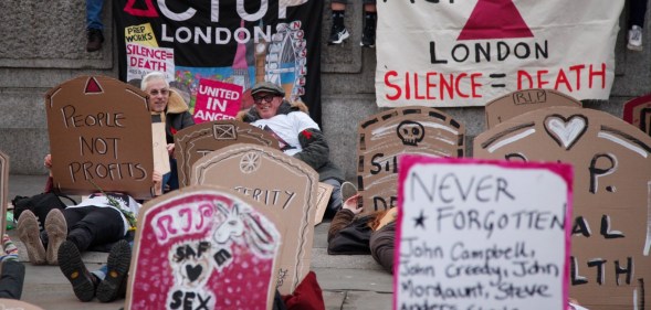 Activists lie down in Trafalgar Square holding cardboard gravestones with messages saying "cuts = coffins", and "queer people are dying" on World AIDS Day