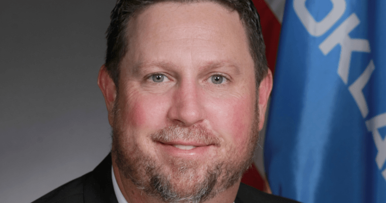 David Bullard, posing for his official US office photo, smiles in front of a set of flags.