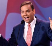 Photo of US congressman George Santos wearing a navy suit, white shirt and pink tie as he makes air quotation gestures with both of his hands.