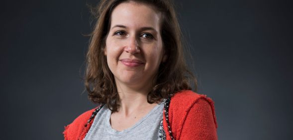 Hadley Freeman, in an orange jumper and grey top, smiles infront of a grey backdrop.