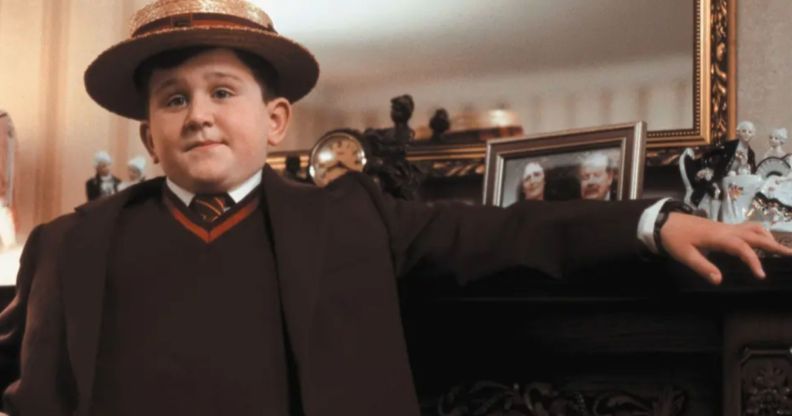 Harry Melling wears a red British school uniform complete with a straw cap and resting his arm against a fireplace while portraying Dudley Dursley in the Harry Potter franchise