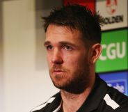 A photo shows Former Australian Football League (AFL) star Dane Swan wearing a black and white top sitting in a room with the names of football sponsors on the wall behind him