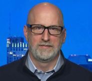 A close-up photo of attorney David French wearing a black top over a blue shirt sitting by a window which has skyscrapers in the background