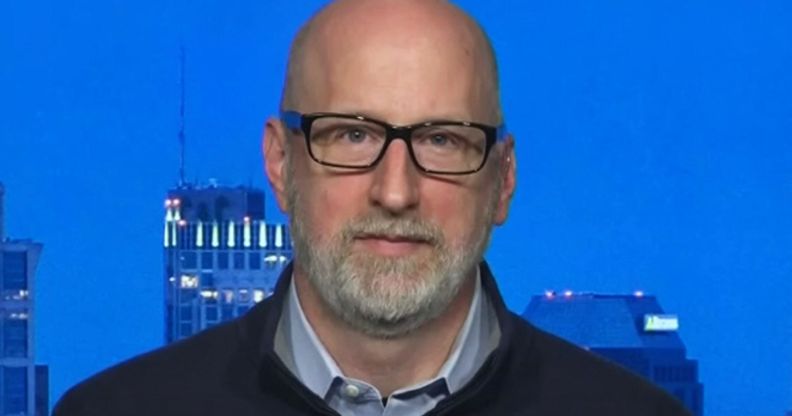 A close-up photo of attorney David French wearing a black top over a blue shirt sitting by a window which has skyscrapers in the background