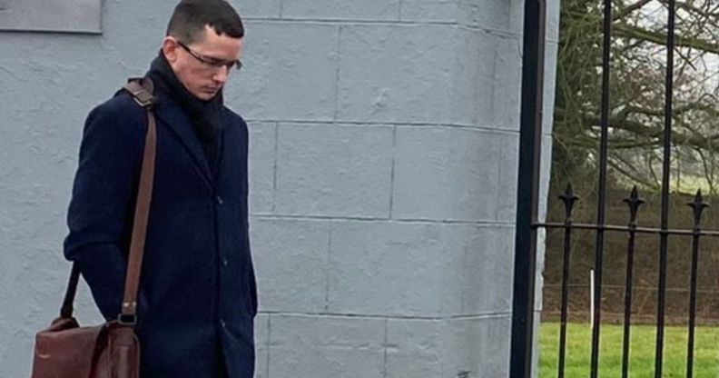 Enoch Burke stands beside a grey school gate, looking down on the floor.