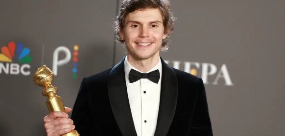 Evan Peters wearing a black suit jacket and bow tie holding his Golden Globe award and standing against a black background with the NBC logo on it