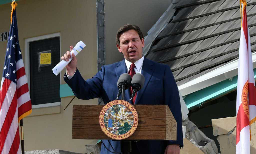 A photo of Florida governor Ron DeSantis wearing a navy suit, white shirt and red tie speaking at a podium