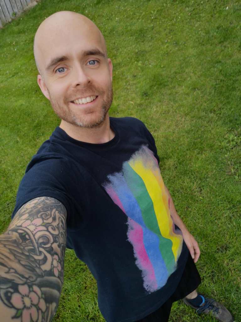 Garry Adair-Gilliland pictured outdoors wearing a black t-shirt with a colourful pattern on the front. The picture is a selfie and his arm is covered in tattoos. Grass is visible in the background.