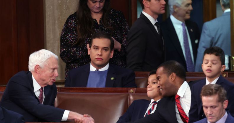 A photo shows US Republican George Santos wearing a dartk suit jacket over a white shirt and light blue sweater sitting alone in the House Chamber of the US Capitol building. All around him you can see other officials looking busy and talking to each other