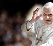 A photo of Pope Benedict XVI looking towards the camera and holding his hand up as if he's making a blessing