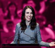 Jacinda Ardern is pictured standing at a podium speaking wearing a grey suit. The background is an edited pink one to show her LGBTQ+ support.