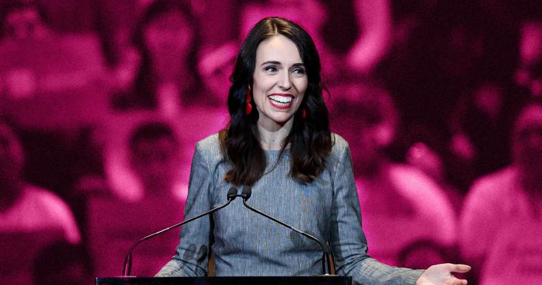 Jacinda Ardern is pictured standing at a podium speaking wearing a grey suit. The background is an edited pink one to show her LGBTQ+ support.