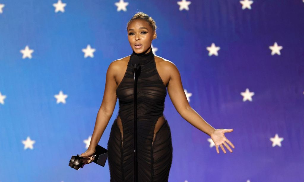 Janelle Monae in a black lace gown holding her Critics Choice Awards, stood in front of a black microphone and against a blue background with white stars on it