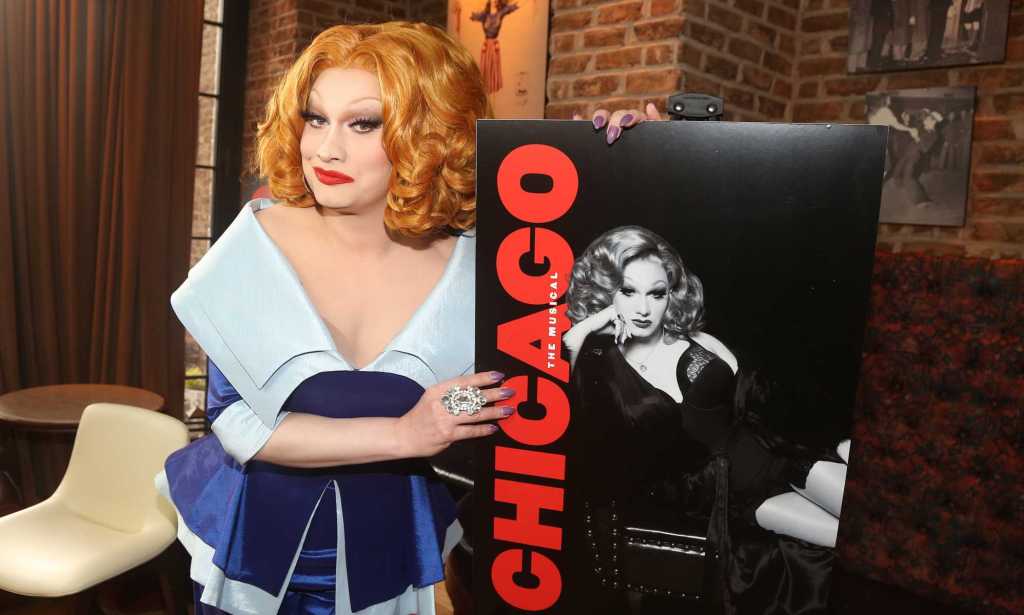 A promo photo of drag queen Jinkx Monsoon wearing a dark and light blue dress holding a poster for the Broadway production of Chicago