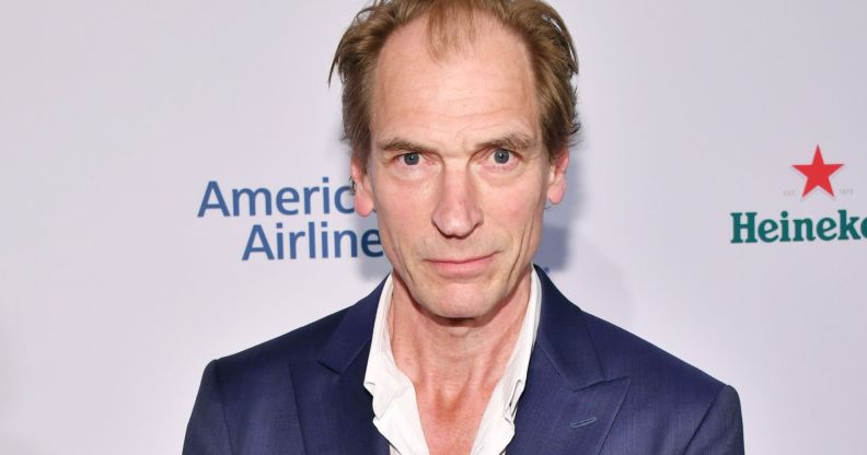 Julian Sands, infront of a white background, smiles on the red carpet.