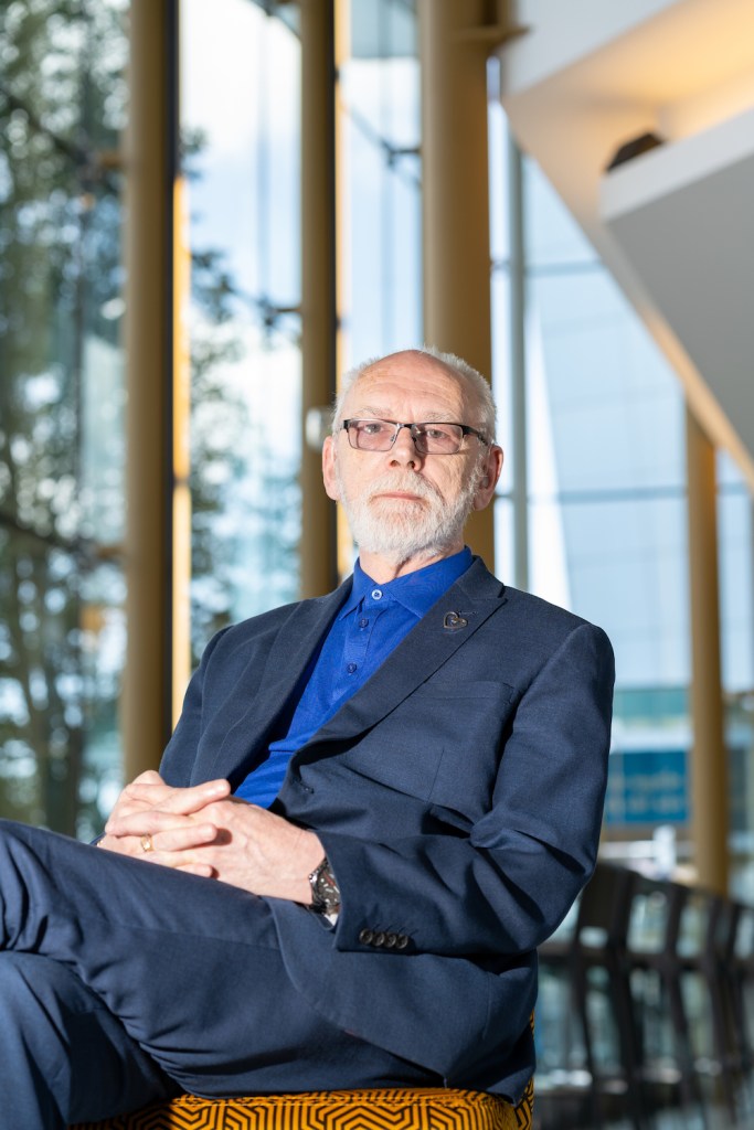 Martyn Butler wearing a dark suit and blue shirt sitting on a chair with windows in the background. He has grey hair and a beard.