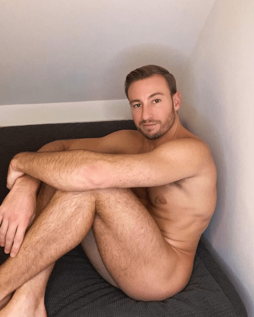 Matthew Mitcham posing nude sitting on a bed.