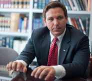 A photo of Florida governor Ron DeSantis wearing a dark suit, white shirt and red tie standing in a room with the background showing books on shelves