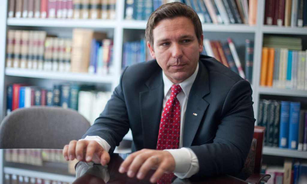 A photo of Florida governor Ron DeSantis wearing a dark suit, white shirt and red tie standing in a room with the background showing books on shelves