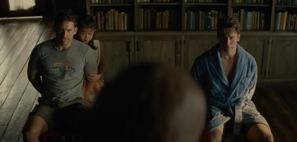 A still from the movie Knock at the Cabin shows two men tied to their chairs in a room with the top half of someone's head looking at them in the foreground of the image