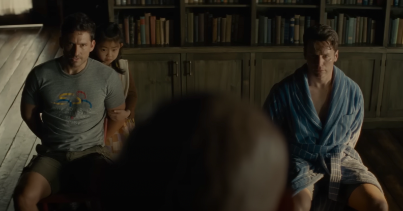 A still from the movie Knock at the Cabin shows two men tied to their chairs in a room with the top half of someone's head looking at them in the foreground of the image