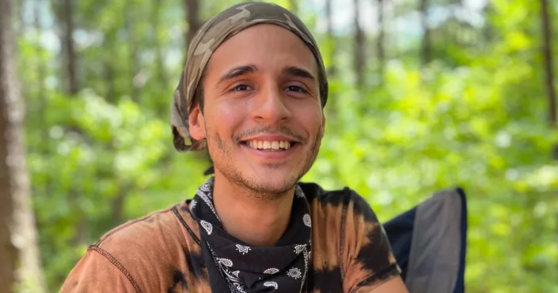 A photo of queer environmental activist Manuel “Tortuguita” Terán wearing a light orange t-shirt and smiling, with a forest in the background