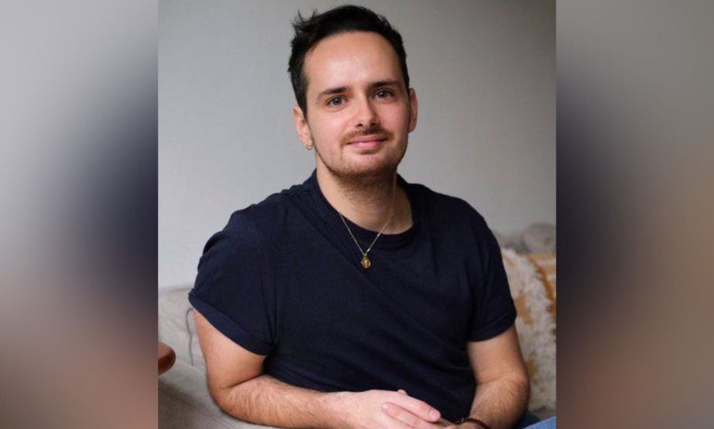 Trans author Harry Nicolas wears a dark top while sitting on a light-coloured sofa with his hands folded in front of him