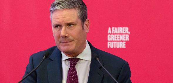 UK Labour Party leader Keir Starmer wears a white button-up shirt, red tie and blue jacket as he speaks at an event. He is standing at a podium in front of a red background