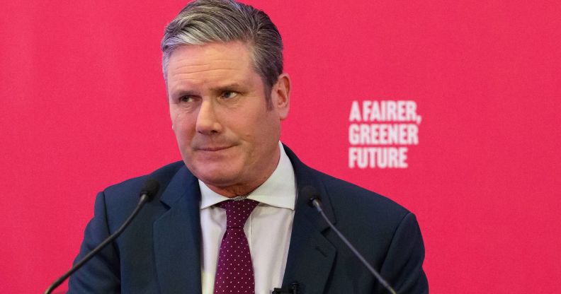 UK Labour Party leader Keir Starmer wears a white button-up shirt, red tie and blue jacket as he speaks at an event. He is standing at a podium in front of a red background