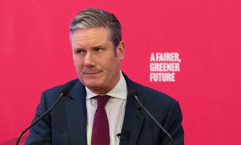 UK Labour Party leader Keir Starmer wears a white button-up shirt, red tie and blue jacket as he speaks at an event. He is standing at a podium in front of a red background